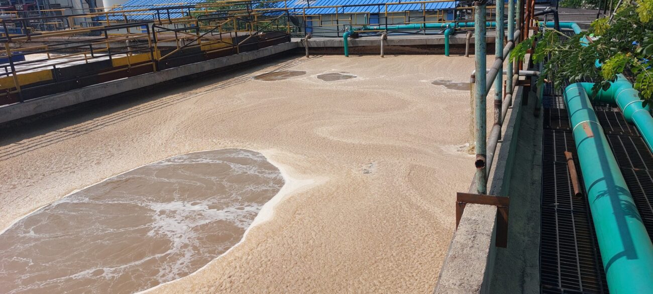 High-angle view of a wastewater treatment basin covered in thick tan biological foam and swirling effluent.