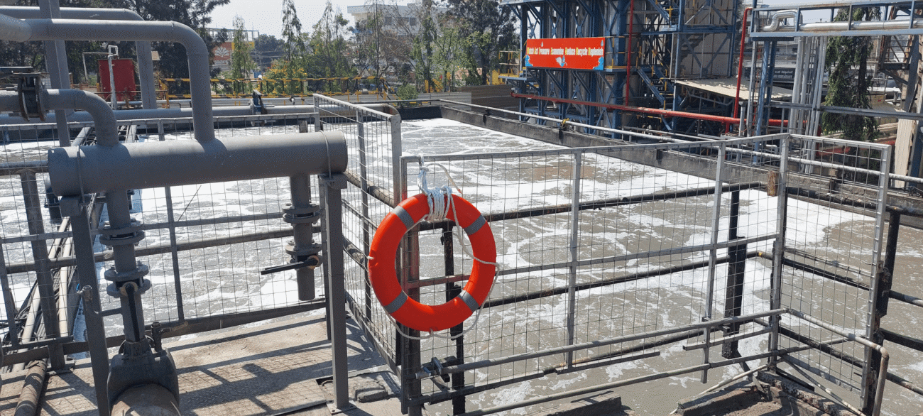 Close-up of an orange rescue lifebuoy mounted to galvanized wire fencing overlooking a vigorously agitated wastewater basin filled with white surface foam, backed by industrial plant equipment.