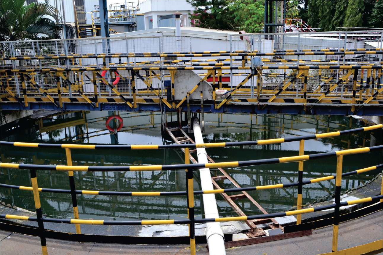 Elevated concrete maintenance catwalk over an active wastewater basin, showing green distribution pipes, valves, pressure gauges, and agitated brown foam to the left.